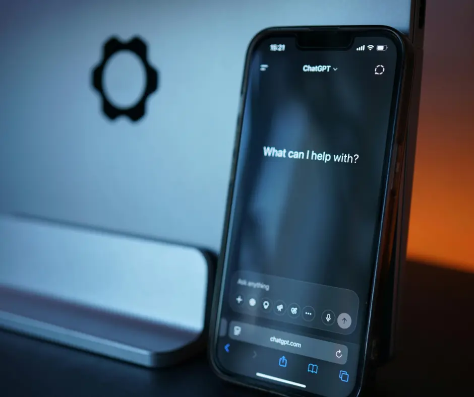 A smartphone mounted on a stand displaying an AI website builder interface with the prompt "What can I help with?" on screen, set against a computer monitor background.